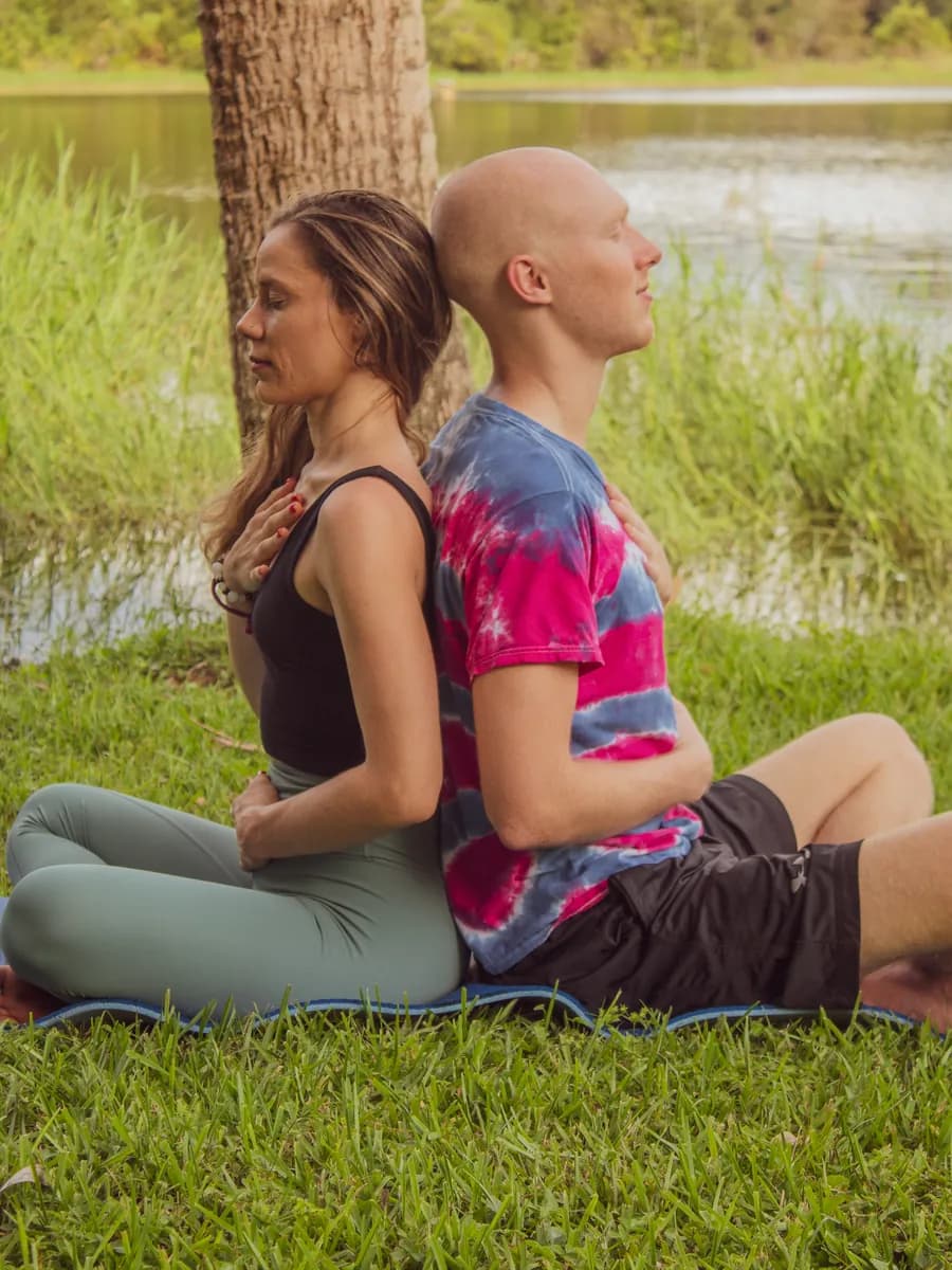 Juliana and Jacob meditating back to back on the grass beside a lake