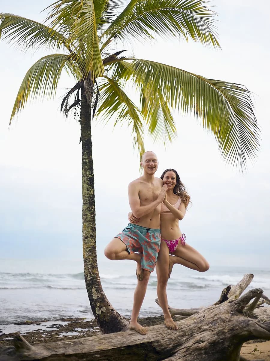 Juliana and Jacob in tree pose together under a palm tree on a beach